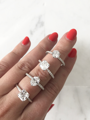Image of a woman wearing four rings, a round solitaire with pave, an Asscher solitiare with pave, and oval solitaire with pave, and an Asscher solitaire all in white gold. Image depicts how our try at home kit helps couples when buying an engagement ring.